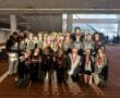 Rowing team celebrating with medals indoors, wearing matching team attire. Smiling group poses for a victorious photo.