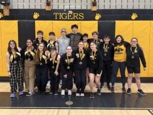 Group of students wearing medals, posing in gym with Tigers sign and yellow-black theme backdrop.
