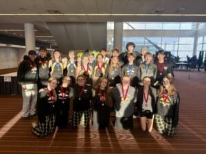 Rowing team celebrating with medals indoors, wearing matching team attire. Smiling group poses for a victorious photo.