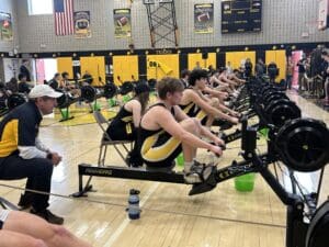 Rowers competing indoors on erg machines, with a coach observing during a school gym event.