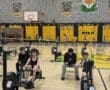 Students compete in a rowing machine race in a school gym with banners and a tiger mascot on the wall.