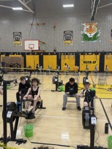 Students compete in a rowing machine race in a school gym with banners and a tiger mascot on the wall.