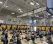 Indoor rowing competition in a gymnasium with athletes on rowing machines, banners, and an American flag visible.