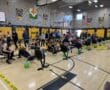 Indoor rowing competition with participants on ergometers in a gymnasium, surrounded by spectators and banners.
