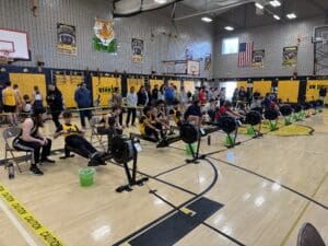 Indoor rowing competition with participants on ergometers in a gymnasium, surrounded by spectators and banners.