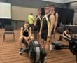 Four athletes in rowing uniforms stand and sit beside indoor rowers at a competition event.