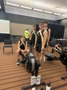 Four athletes in rowing uniforms stand and sit beside indoor rowers at a competition event.