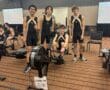 Four young athletes in matching gear stand and sit by rowing machines indoors.