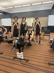 Four young athletes in matching gear stand and sit by rowing machines indoors.
