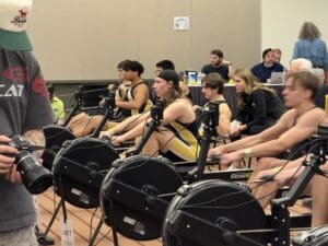 Indoor rowing competition with athletes focused on training, supported by coaches and spectators in the background.