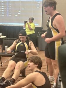 Rowing team in action during an indoor competition, focused on an ergometer with cheering teammates nearby.
