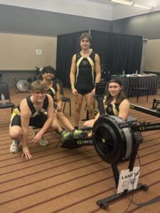 Four athletes in rowing gear pose with an indoor rowing machine in a gym setting.
