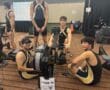 Rowing team preparing for competition on indoor rowing machines, Lane 15, with scoreboard in background.