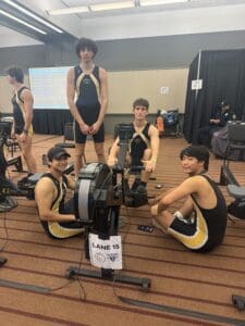 Rowing team preparing for competition on indoor rowing machines, Lane 15, with scoreboard in background.