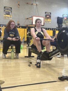 Indoor rowing competition at Marshall Middle School gym, focused athlete on ergometer, coach supervising.