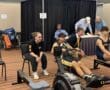 Two athletes rowing on machines, with a coach cheering them on at an indoor event.