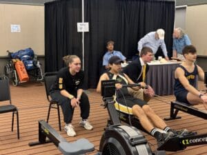 Two athletes rowing on machines, with a coach cheering them on at an indoor event.