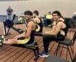 Rowers in yellow and black uniforms on indoor rowing machines, smiling during a competition.