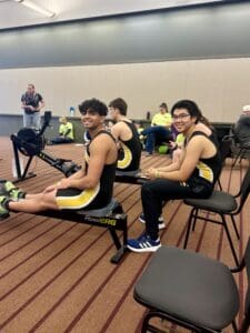 Rowers in yellow and black uniforms on indoor rowing machines, smiling during a competition.