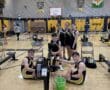 Rowing team poses next to indoor rowing machines in a gymnasium.