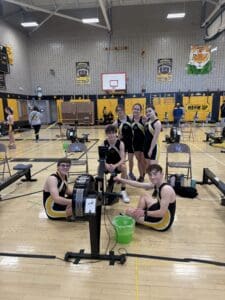 Rowing team poses next to indoor rowing machines in a gymnasium.