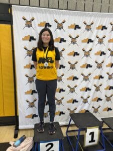 Young athlete on podium wearing yellow Rowing jersey with medal, standing before branded backdrop at sports event.
