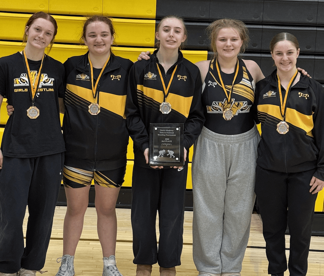 Five female wrestlers with medals and a trophy, wearing team uniforms, stand in a gym for a group photo.