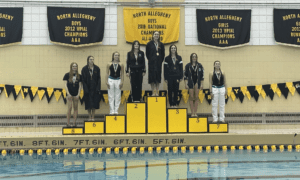 High school swimming medalists on podium at North Allegheny pool with championship banners in the background.
