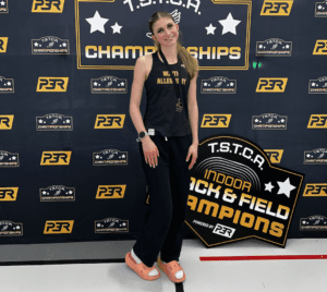 Athlete posing at TSTCA Indoor Track & Field Championships backdrop, wearing casual sportswear.