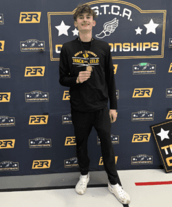 Young athlete holding medal at TSTCA Championships in track and field gear stands smiling against branded backdrop.