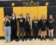 Rowing team members pose in matching attire and medals in a gym with Tigers sign in background.