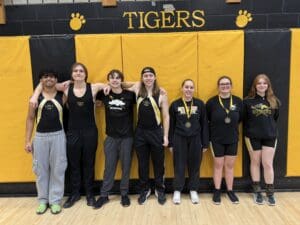 Rowing team members pose in matching attire and medals in a gym with Tigers sign in background.