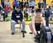 Teen rowing on an indoor machine at Marshall Middle School gym event, with coach and other participants in the background.