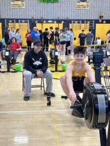 Teen rowing on an indoor machine at Marshall Middle School gym event, with coach and other participants in the background.