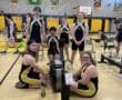 Rowing team posing in gym with rowing machines, wearing black and yellow uniforms, gym banners in background.