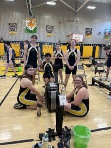 Rowing team posing in gym with rowing machines, wearing black and yellow uniforms, gym banners in background.