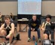 Two athletes rowing indoors with a coach during a competitive training session, screen displaying progress.