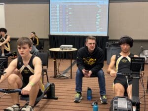 Two athletes rowing indoors with a coach during a competitive training session, screen displaying progress.