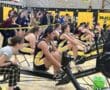 Rowing team competes indoors on rowing machines, with supporters cheering enthusiastically in a gymnasium setting.