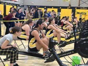Rowing team competes indoors on rowing machines, with supporters cheering enthusiastically in a gymnasium setting.