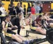 Rowers compete on indoor rowing machines in a gym, with spectators watching closely in the background.