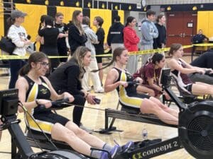 Rowers compete on indoor rowing machines in a gym, with spectators watching closely in the background.