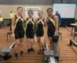 Four women in matching athletic uniforms at a rowing championship event, standing beside indoor rowing machines.