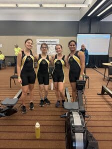 Four women in matching athletic uniforms at a rowing championship event, standing beside indoor rowing machines.