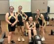 Four women in rowing uniforms smiling beside an ergometer in an indoor setting, competition results displayed on screen.