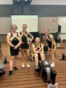 Four women in rowing uniforms smiling beside an ergometer in an indoor setting, competition results displayed on screen.