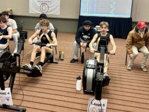 Two athletes rowing on ergometers at an indoor competition with a scoreboard in the background.