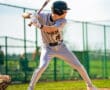 A youth baseball player in a grey uniform prepares to swing a bat on a sunny day at the ballpark.