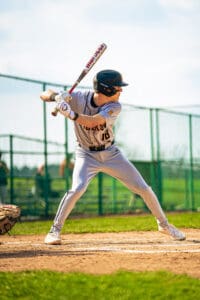 A youth baseball player in a grey uniform prepares to swing a bat on a sunny day at the ballpark.