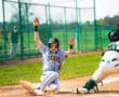 Baseball player sliding into home plate with hands raised, wearing a Tigers jersey, during a sunny game day.